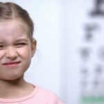 Dziewczynka mrużąca oczy podczas badania wzroku – w tle tablica okulistyczna, możliwy problem z ostrością widzenia.