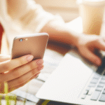 Osoba pracująca przy laptopie, trzymająca w jednej ręce smartfon, a drugą wpisująca coś na klawiaturze. W tle widoczny kubek i rozmazane książki