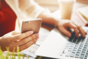 Osoba pracująca przy laptopie, trzymająca w jednej ręce smartfon, a drugą wpisująca coś na klawiaturze. W tle widoczny kubek i rozmazane książki