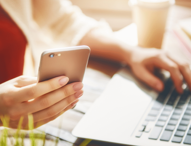 Osoba pracująca przy laptopie, trzymająca w jednej ręce smartfon, a drugą wpisująca coś na klawiaturze. W tle widoczny kubek i rozmazane książki
