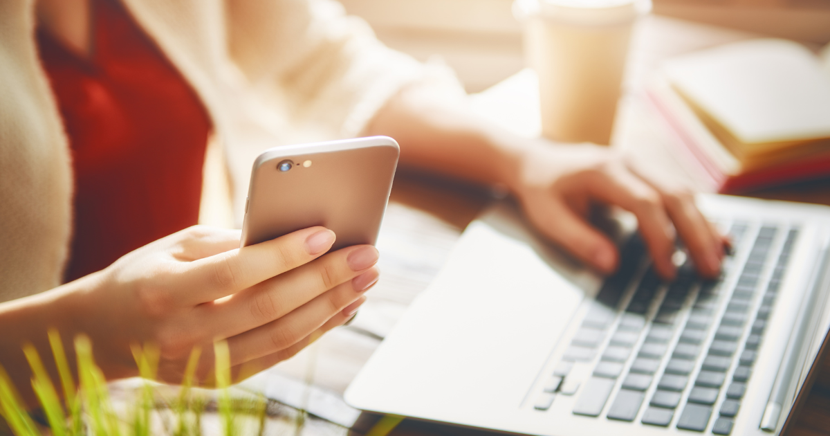 Osoba pracująca przy laptopie, trzymająca w jednej ręce smartfon, a drugą wpisująca coś na klawiaturze. W tle widoczny kubek i rozmazane książki