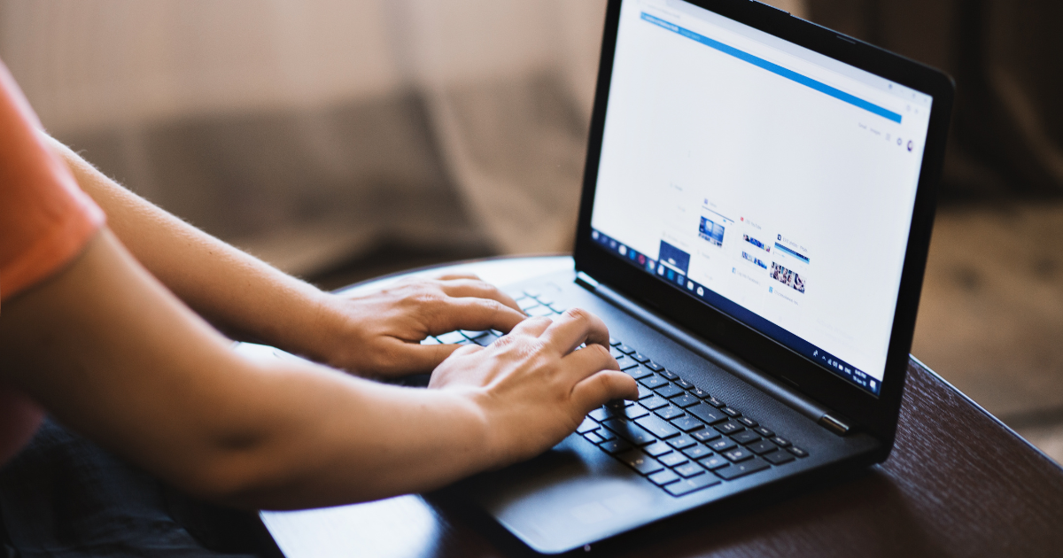 Osoba korzystająca z laptopa, siedząca przy stole i wpisująca coś na klawiaturze. Na ekranie widać otwartą przeglądarkę internetową