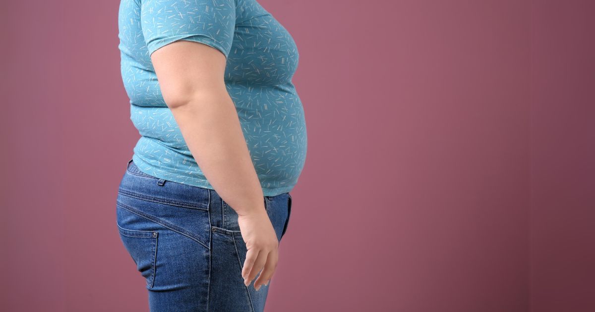 Osoba z nadwagą w pozycji bocznej na tle różowej ściany, widoczny brzuch i biodra