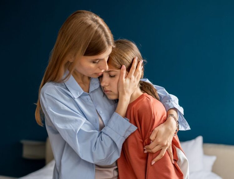 Zatroskana matka przytula smutną córkę, próbując ją pocieszyć – scena ukazująca wsparcie emocjonalne i bliską więź w trudnej chwili