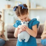 Mała dziewczynka tuląca pluszowego misia stoi ze smutną miną, w tle jej rodzice siedzą odwróceni od siebie, wyraźnie skłóceni