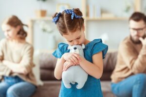 Mała dziewczynka tuląca pluszowego misia stoi ze smutną miną, w tle jej rodzice siedzą odwróceni od siebie, wyraźnie skłóceni