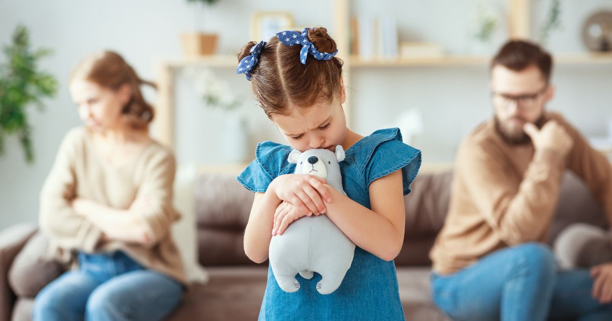 Mała dziewczynka tuląca pluszowego misia stoi ze smutną miną, w tle jej rodzice siedzą odwróceni od siebie, wyraźnie skłóceni