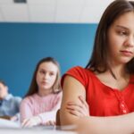 Smutna nastolatka w czerwonej koszulce siedzi w ławce szkolnej z założonymi rękami, podczas gdy w tle inni uczniowie patrzą w jej stronę z obojętnością lub dezaprobatą, co sugeruje sytuację wykluczenia lub izolacji w klasie