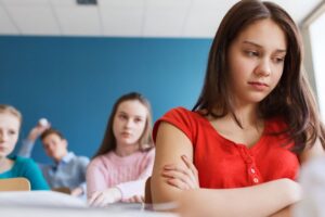 Smutna nastolatka w czerwonej koszulce siedzi w ławce szkolnej z założonymi rękami, podczas gdy w tle inni uczniowie patrzą w jej stronę z obojętnością lub dezaprobatą, co sugeruje sytuację wykluczenia lub izolacji w klasie