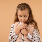 Mała dziewczynka o długich, brązowych włosach patrzy w dół, trzymając kurteczkę lub kamizelkę blisko klatki piersiowej, na tle jednolitej, beżowej ściany