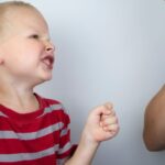 Mały chłopiec w czerwono-szarej koszulce w paski z zaciśniętymi pięściami i gniewnym wyrazem twarzy grozi uderzeniem starszej osoby, która zasłania usta ręką